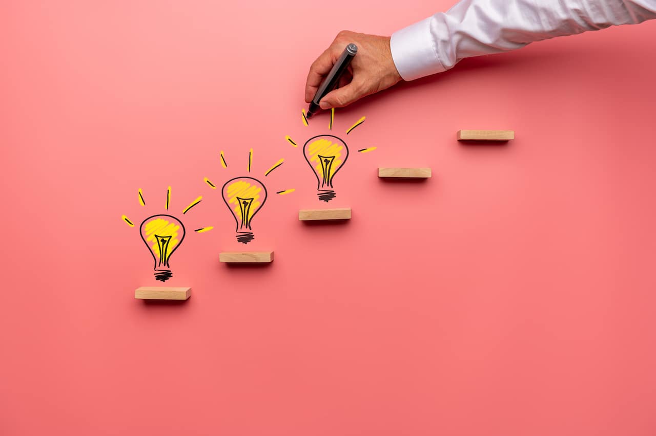 Male hand drawing yellow light bulbs on wooden steps in a conceptual image. Over pink background.